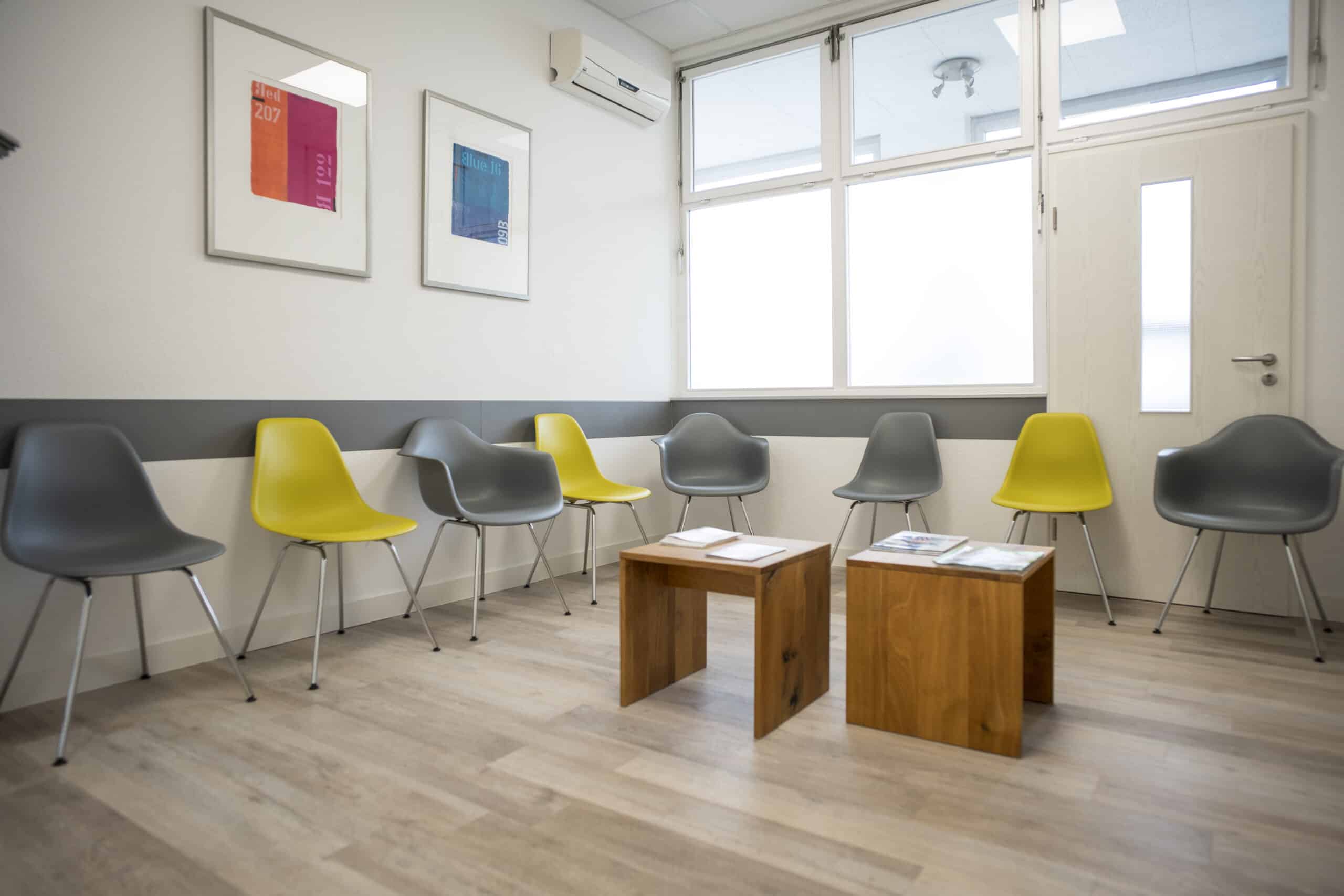 Helles Wartezimmer mit Stühlen in Grau und Gelb, freundliche Atmosphäre, Wartebereich mit Magazinen.